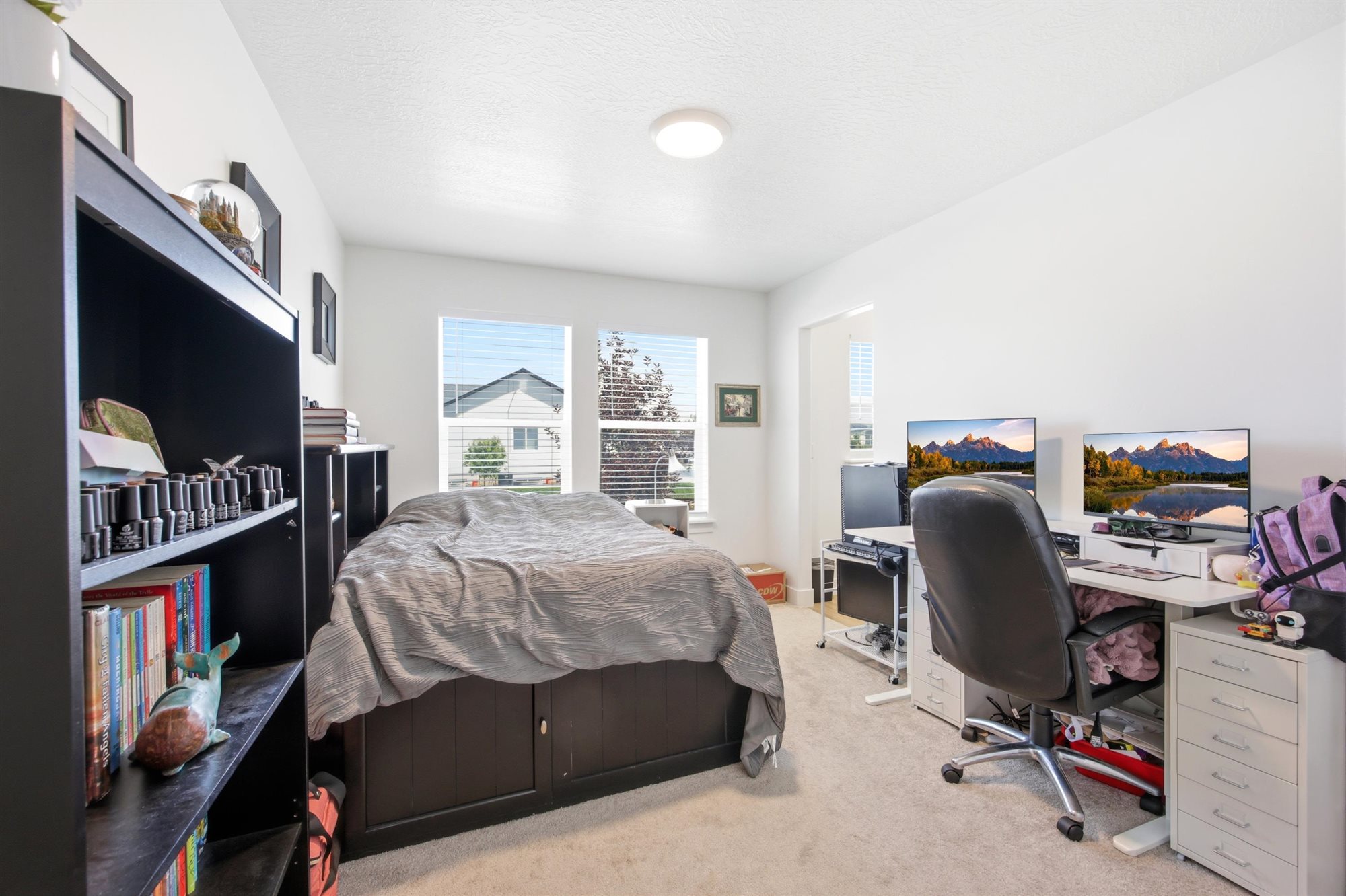 Carpeted room with white walls, featuring a built-in dark wood bookshelf, a bed with integrated storage, and two windows with blinds