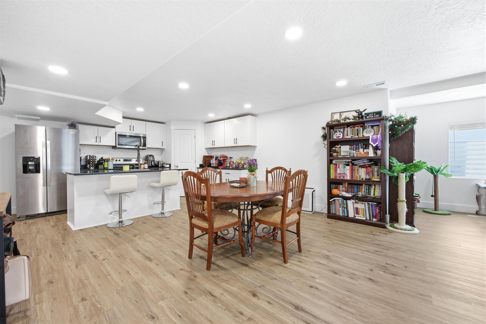 Open-concept living space featuring wood-finish flooring, recessed lighting, and a kitchen with stainless steel appliances, white cabinetry, and a black countertop island with seating