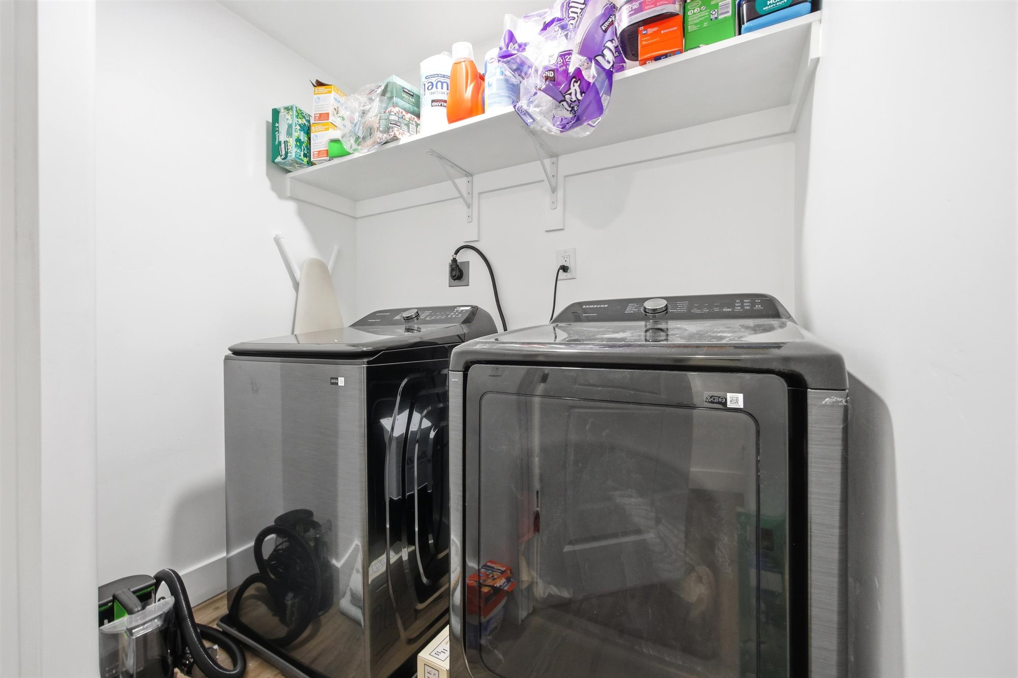 Dedicated utility area featuring white walls, wood-finish flooring, a wall-mounted white storage shelf, and electrical outlets