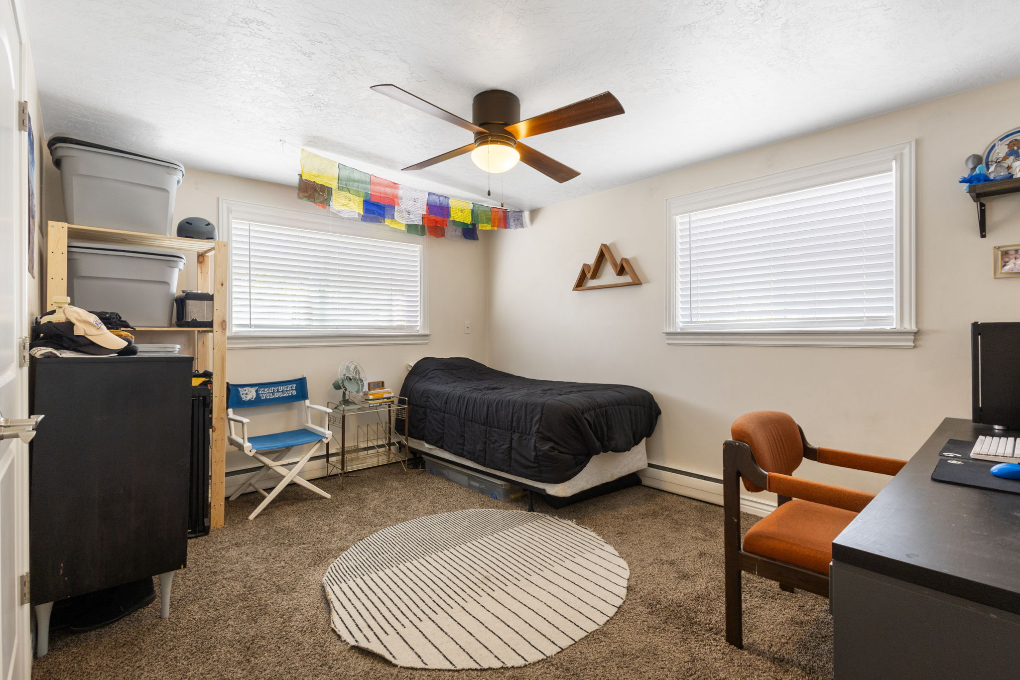 Carpeted interior space featuring dual windows with blinds, a ceiling fan with integrated lighting, and white baseboards