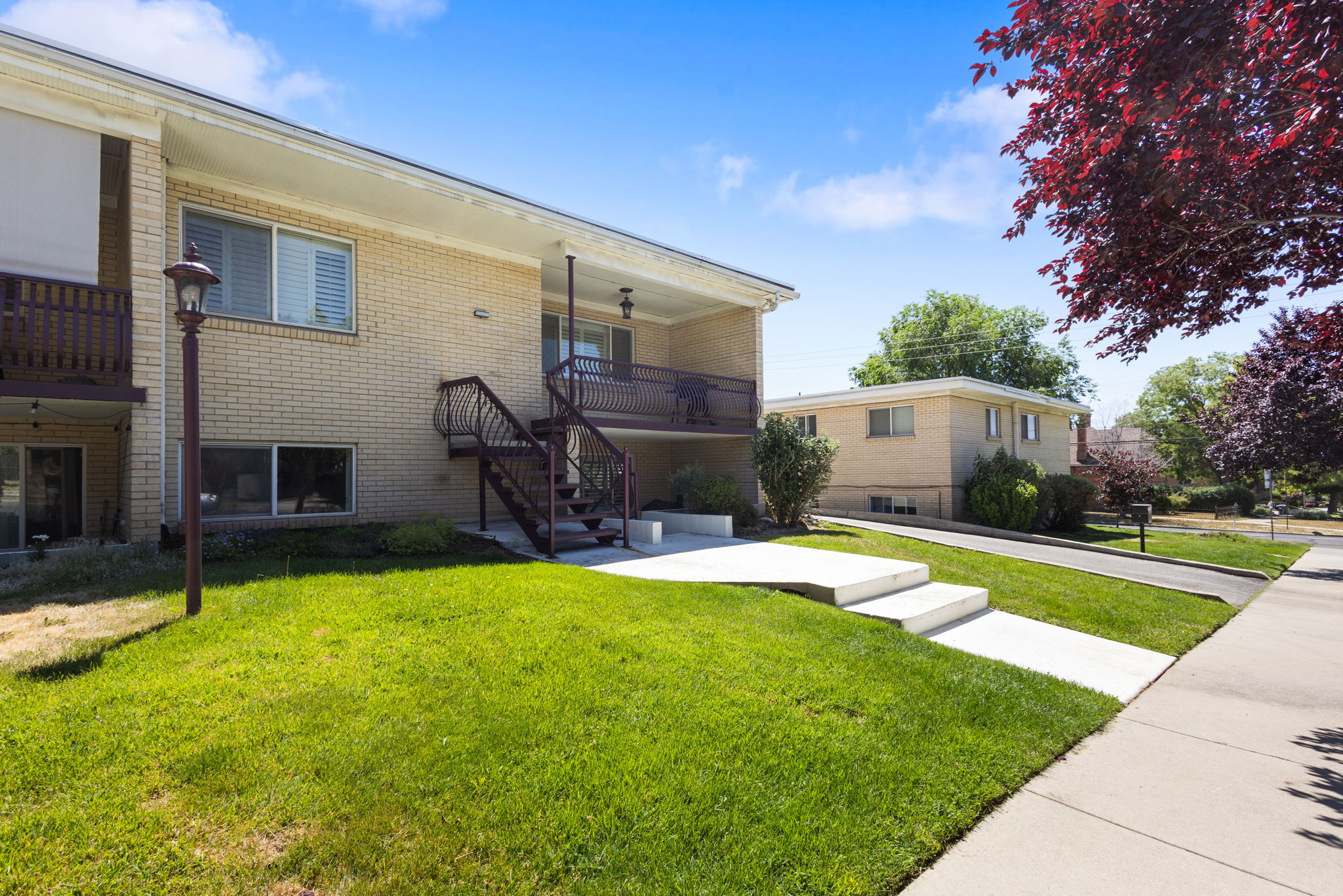 Brick multi-unit building featuring balconies with dark railings, a prominent exterior staircase, and mature landscaping