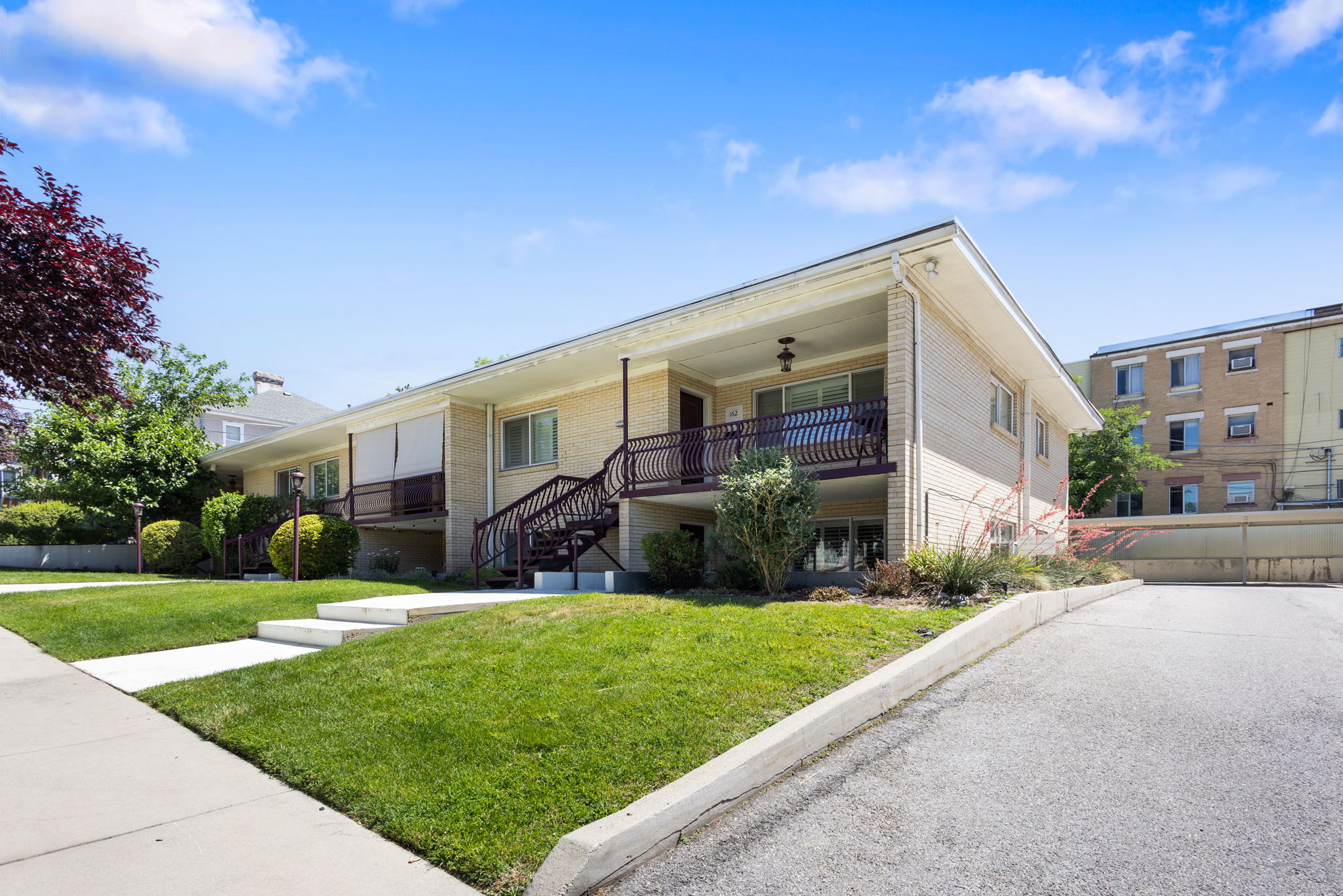 Multi-unit building featuring a light brick exterior, manicured lawn, concrete walkways, and an asphalt driveway