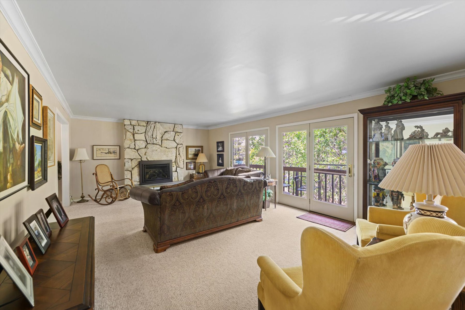 Living room featuring a stone-clad fireplace, light-colored carpeting, crown molding, and double glass doors opening to a balcony with black railings
