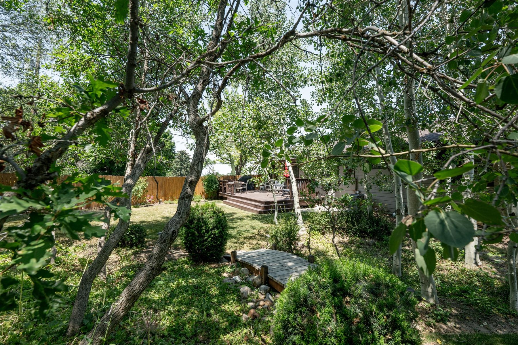Lush backyard featuring mature trees, a multi-level deck, fenced perimeter, and a stone footbridge