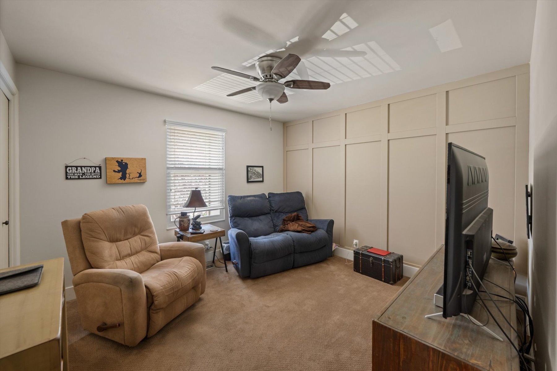 Room featuring a beige accent wall with panel molding, neutral carpet flooring, a window with blinds, and a ceiling fan with integrated lighting