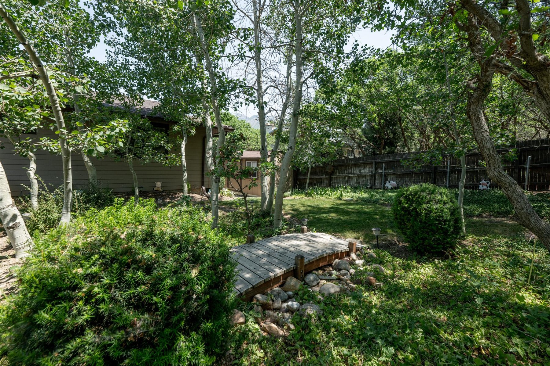 Expansive backyard featuring a curved wooden bridge over a rock bed, mature trees, manicured shrubs, and a partial view of the home's exterior with horizontal siding