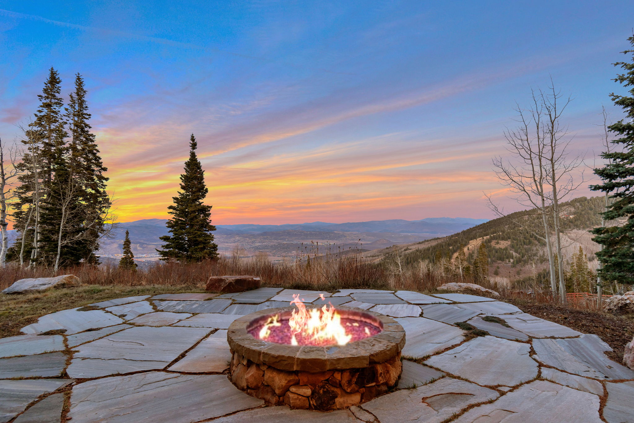 Stone patio featuring a built-in fire pit with stone surround, surrounded by mature evergreen trees
