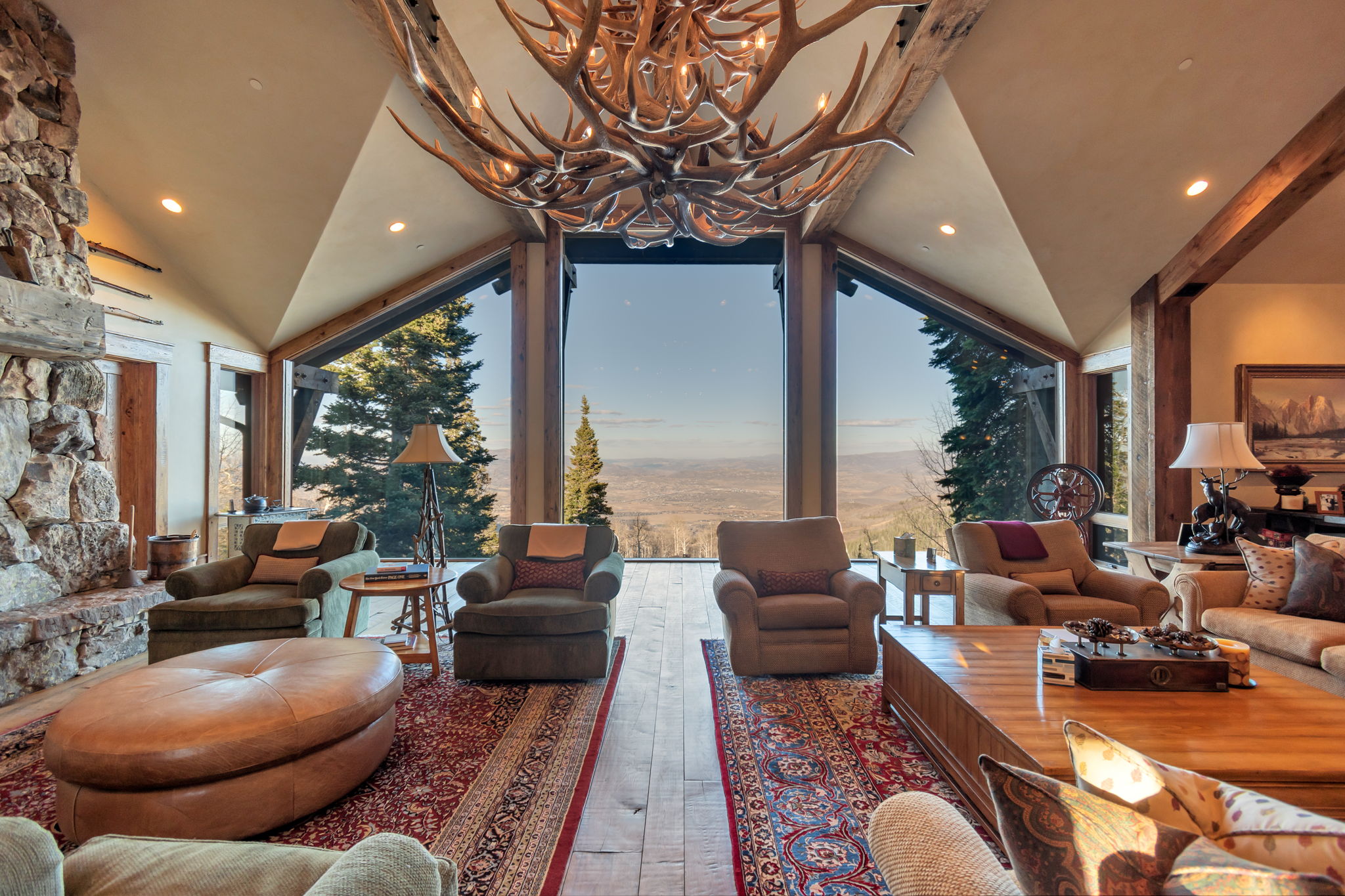 Vaulted living space featuring a large antler chandelier, exposed wood beams, a stone fireplace, and a wall of windows