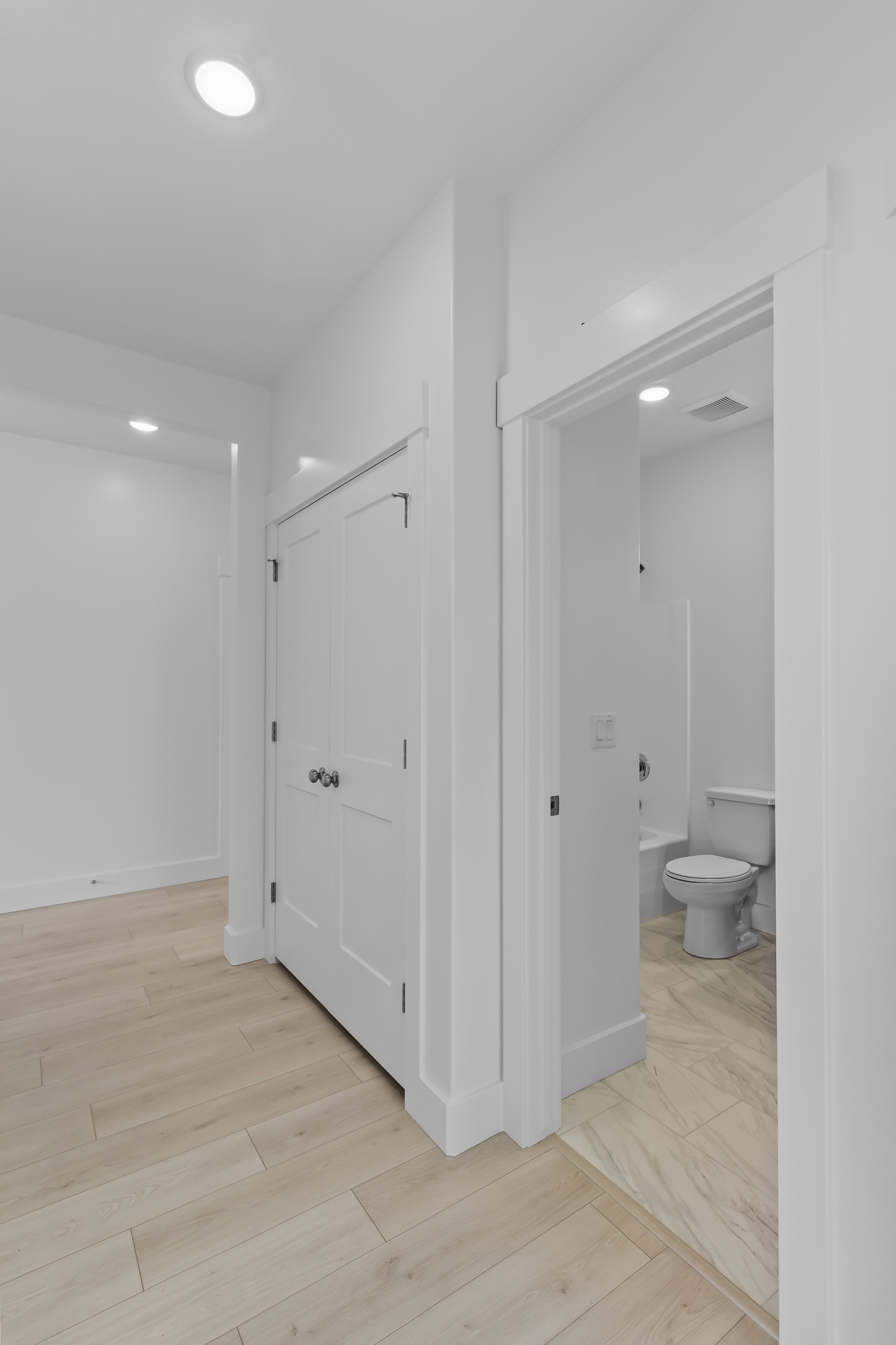 Light wood-finish flooring extending into a white-trimmed doorway, revealing a bathroom with a white toilet and tub