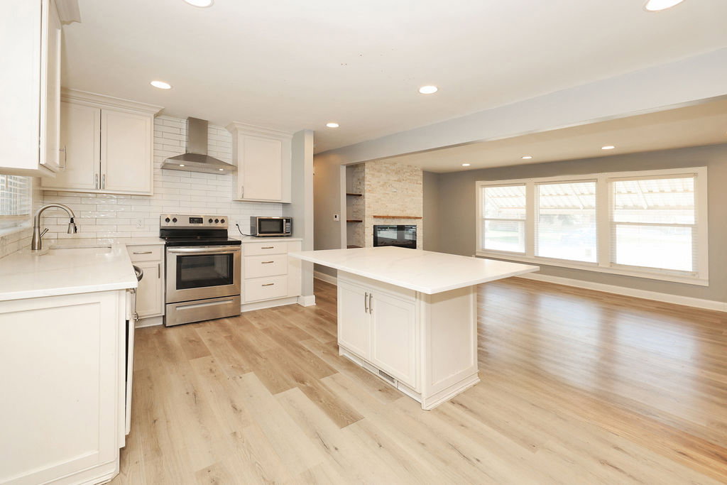 Open-concept kitchen featuring an island with a light-toned countertop, white cabinetry, stainless steel appliances, and wood-finish flooring extending into the adjacent living area with large windows