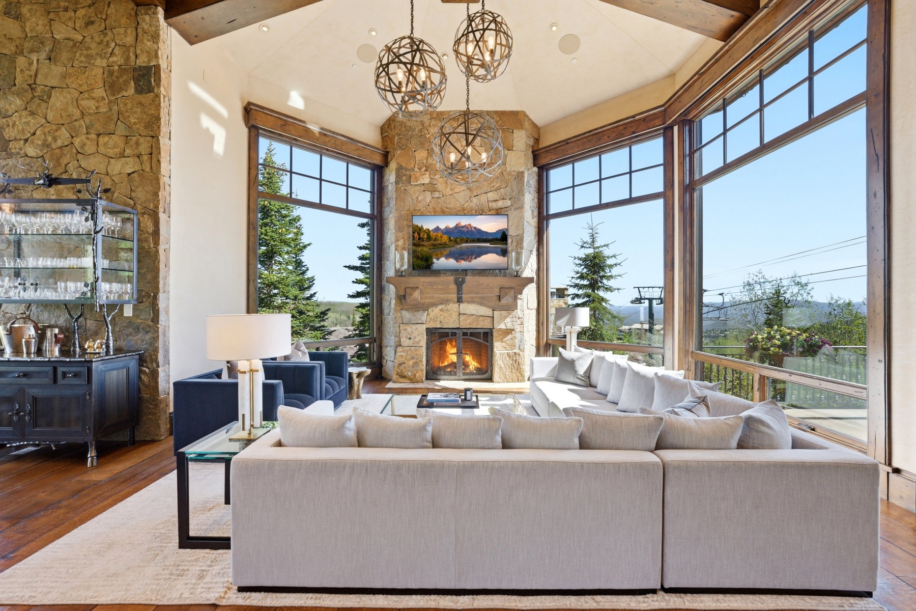 Vaulted great room featuring expansive windows, a stone-clad fireplace, exposed wood beams, and warm wood-finish flooring