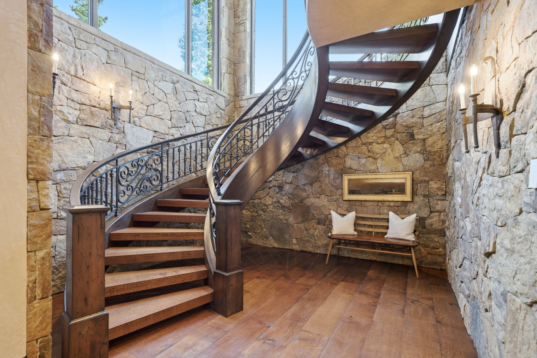 Curved wooden staircase with ornate wrought iron railing, stone wall finishes, wood-finish flooring, and integrated wall sconce lighting
