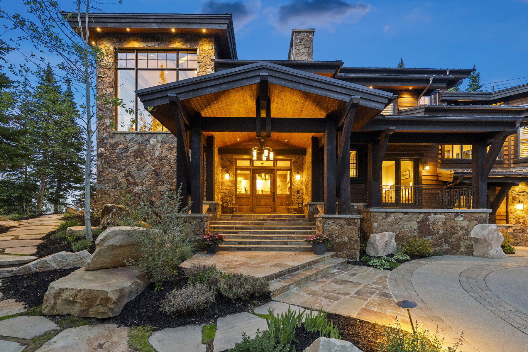 Stone and timber-frame exterior featuring a grand covered entry with wood double doors, multiple large windows, a stone chimney, and a stone slab walkway