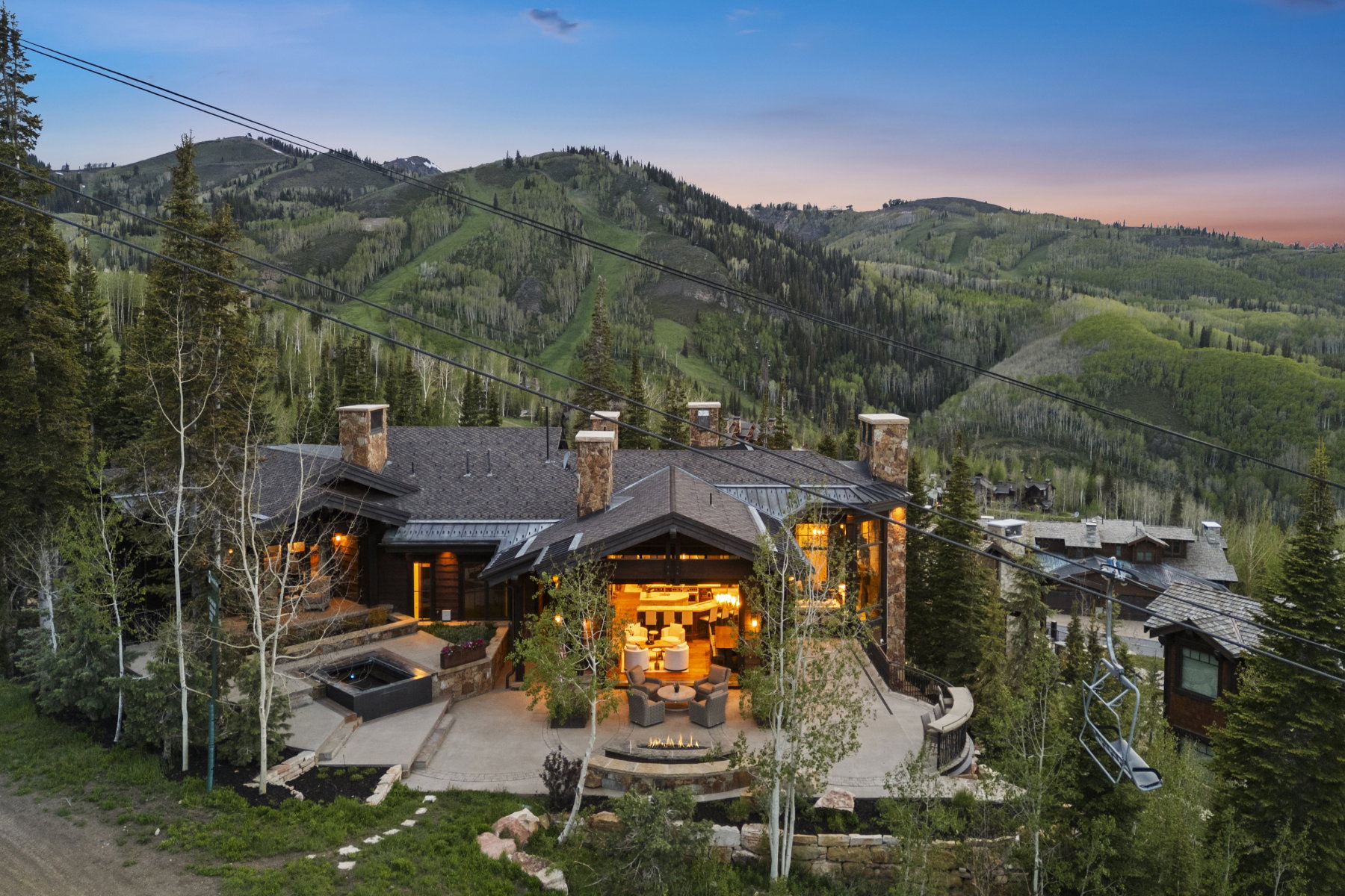 Mountain-style residence featuring multiple stone chimneys, a large outdoor patio with a built-in fire pit, an integrated spa, and a dark shingled roof