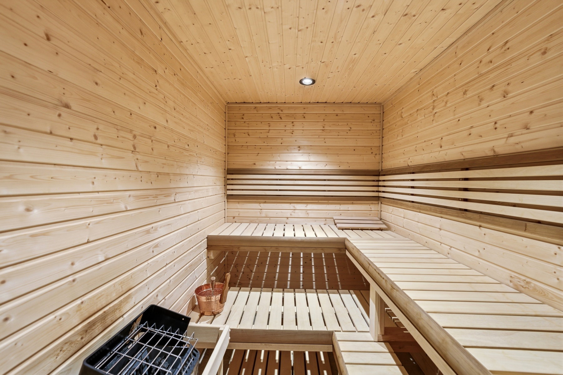 Cedar-paneled sauna featuring a built-in multi-level bench, recessed ceiling lighting, and a floor-mounted heater