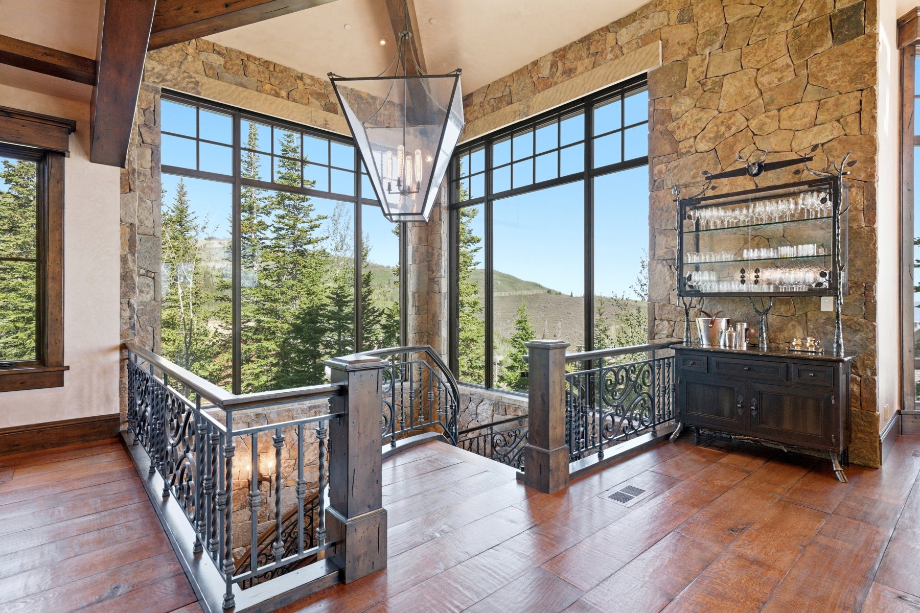 Expansive floor-to-ceiling windows with black framing, wood-finish flooring, stone accent wall, decorative metal balustrade, and exposed wood beam ceiling