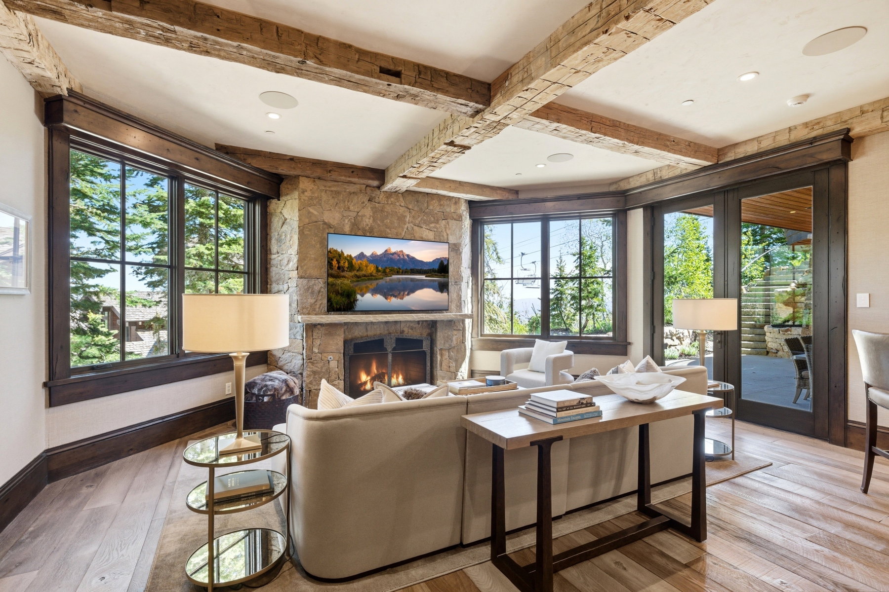 Living area featuring exposed wood beams, a stone-clad fireplace with a hearth, wood-finish flooring, and multiple large windows with dark wood trim