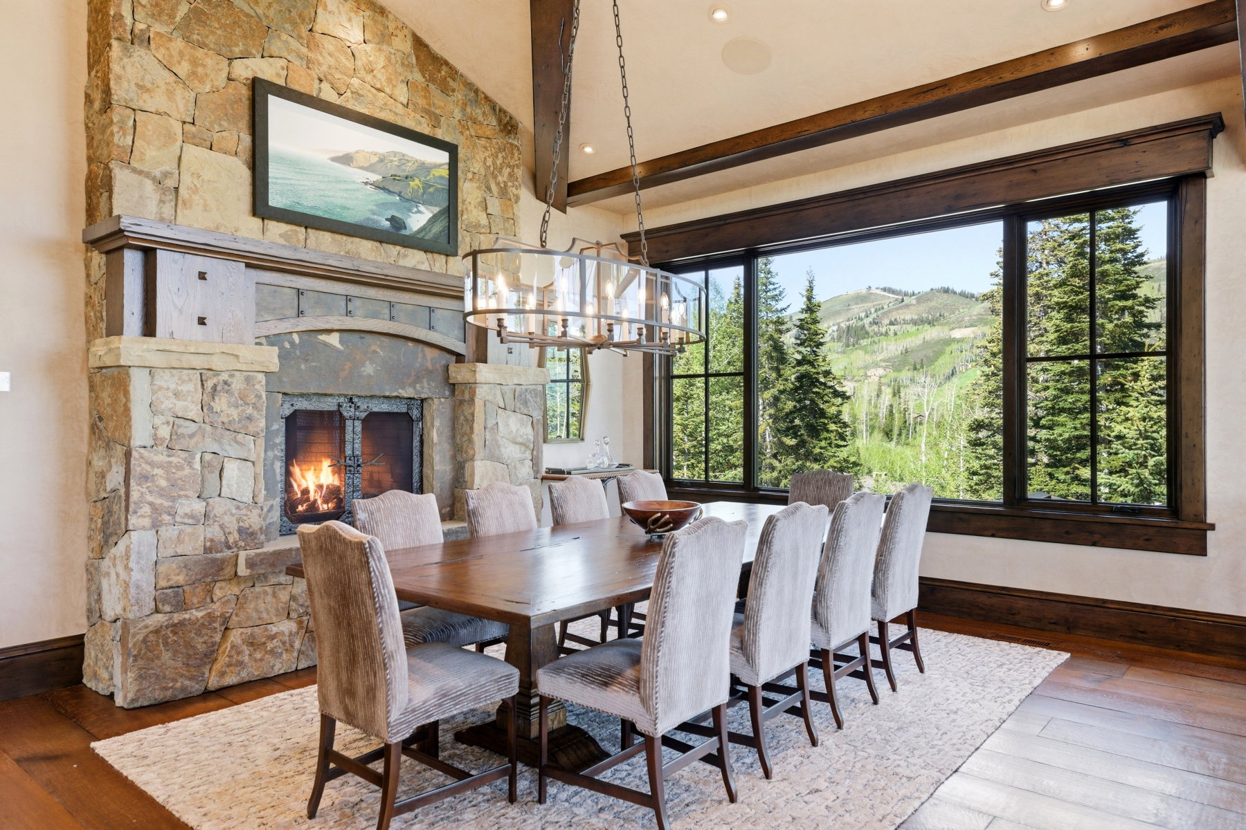 Stone fireplace with wood mantel, a large window with outdoor scenery, recessed lighting, wood-finish flooring, and exposed wood beams