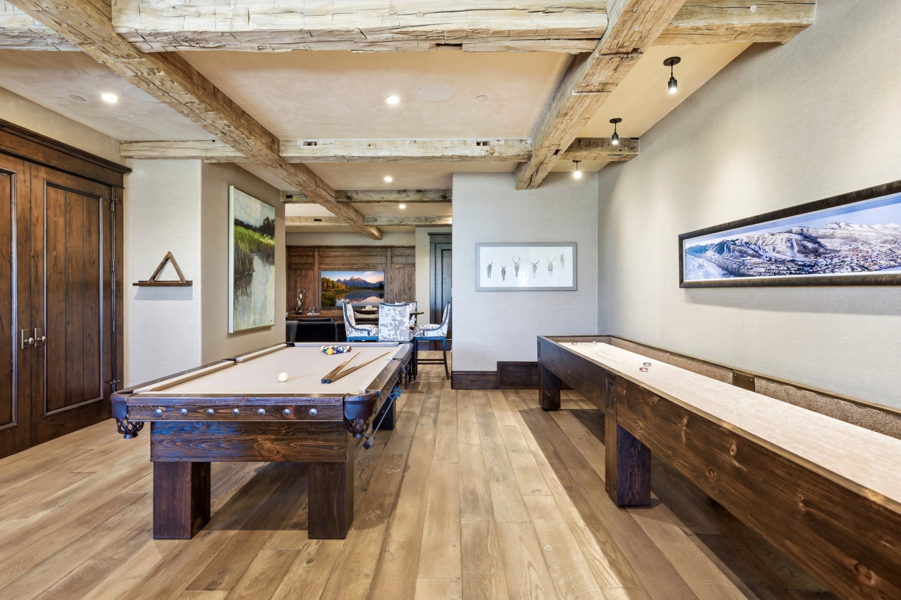 Spacious recreation room featuring exposed wood ceiling beams, light wood-finish flooring, recessed lighting, and dark wood double doors
