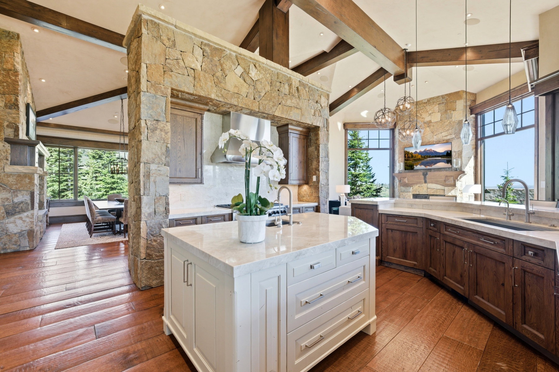 Gourmet kitchen featuring a substantial central island, stainless steel appliances, wood-finish flooring, exposed ceiling beams, and stone wall accents