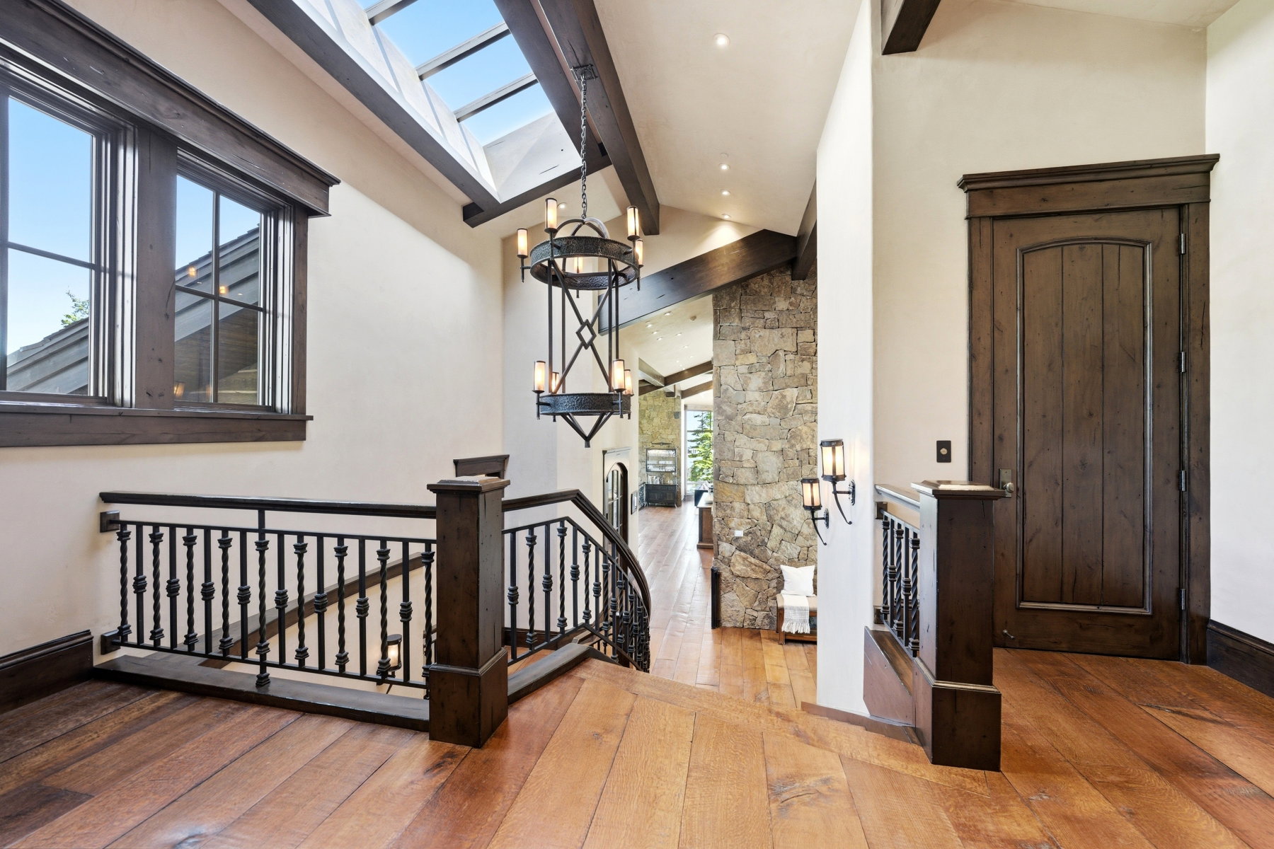 Hardwood flooring throughout a spacious hallway featuring a wrought iron staircase railing with dark wood posts, a wood paneled door, and a skylight