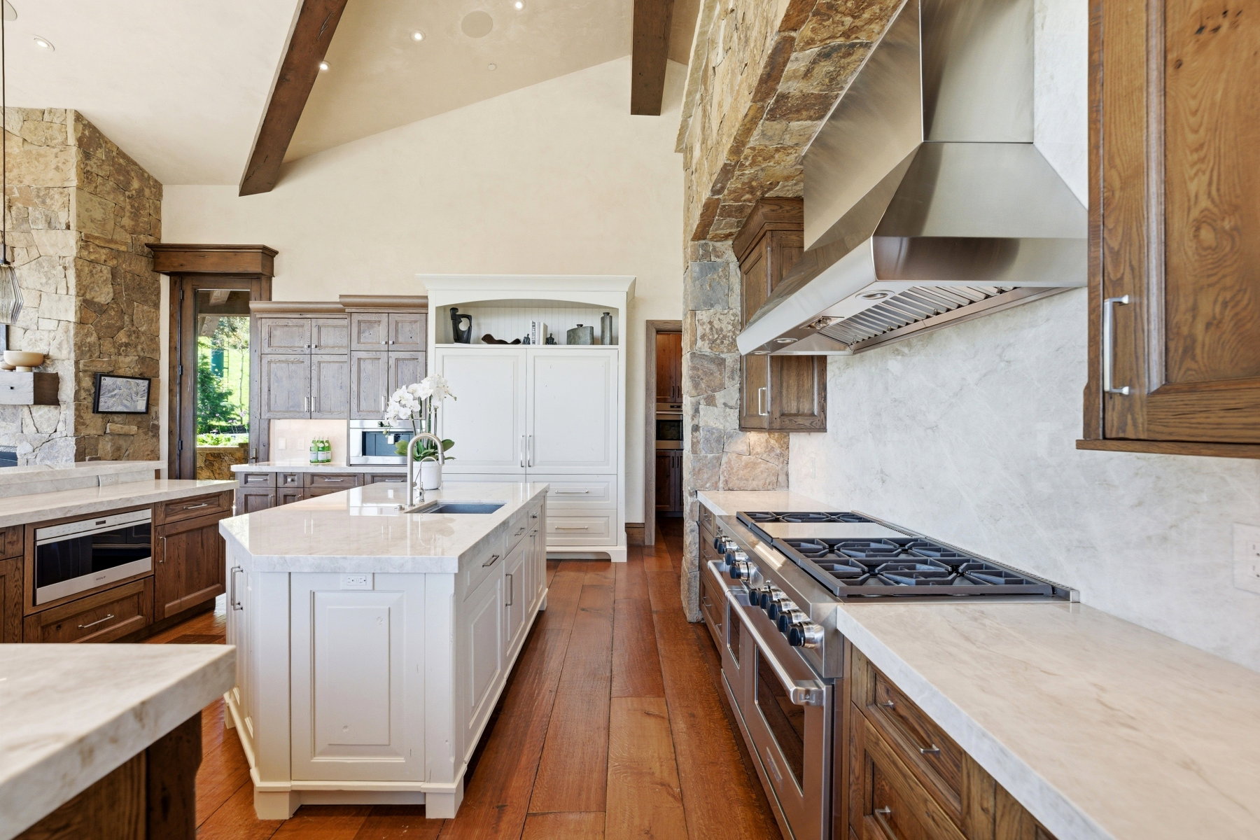Gourmet kitchen featuring a large central island with an undermount sink, stainless steel appliances, varied wood cabinetry, stone accent walls, and vaulted ceilings with exposed beams