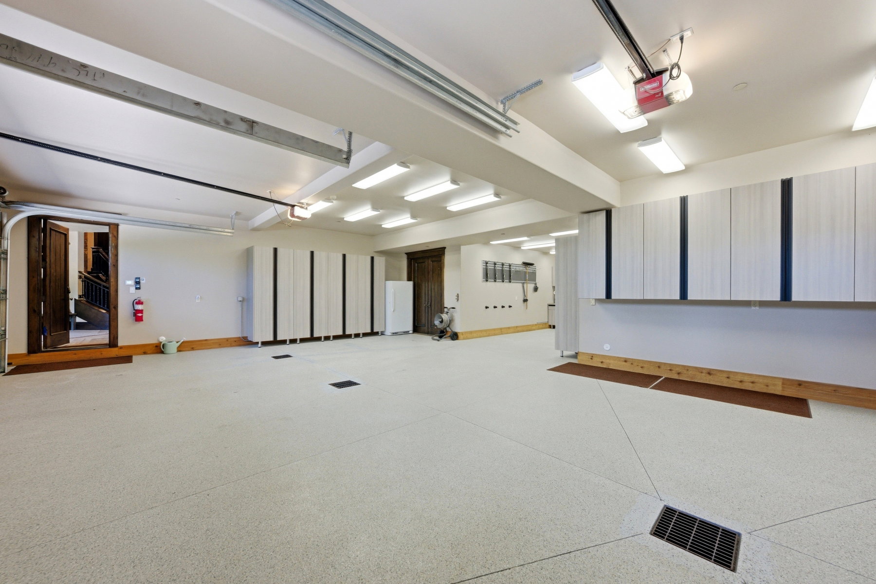 Spacious garage featuring a light-colored epoxy floor with integrated drains, extensive built-in storage cabinetry, overhead fluorescent lighting, and a dark wood entry door