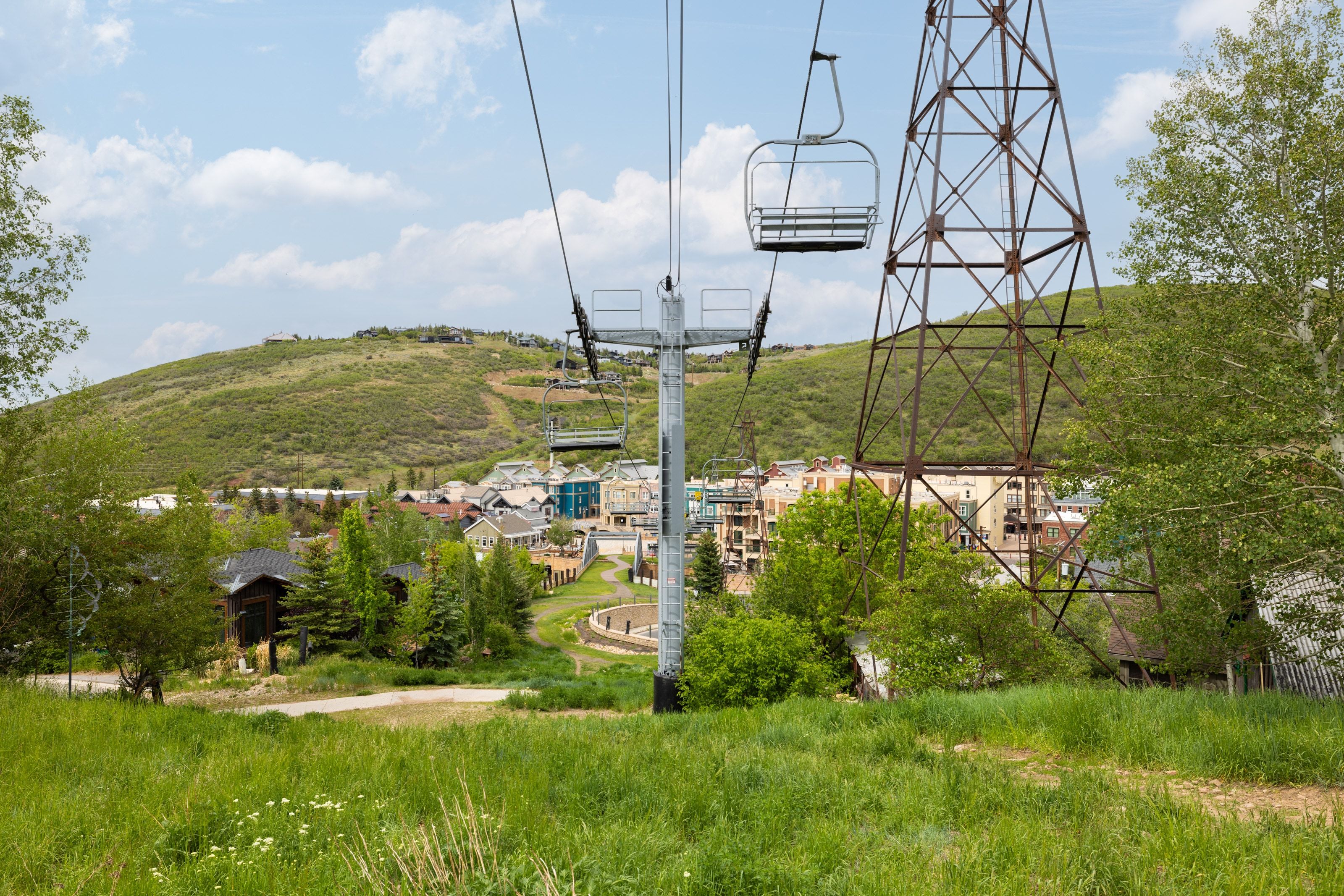 Lush green hillside with a ski lift structure, residential buildings, and distant mountainous terrain under a partly cloudy sky