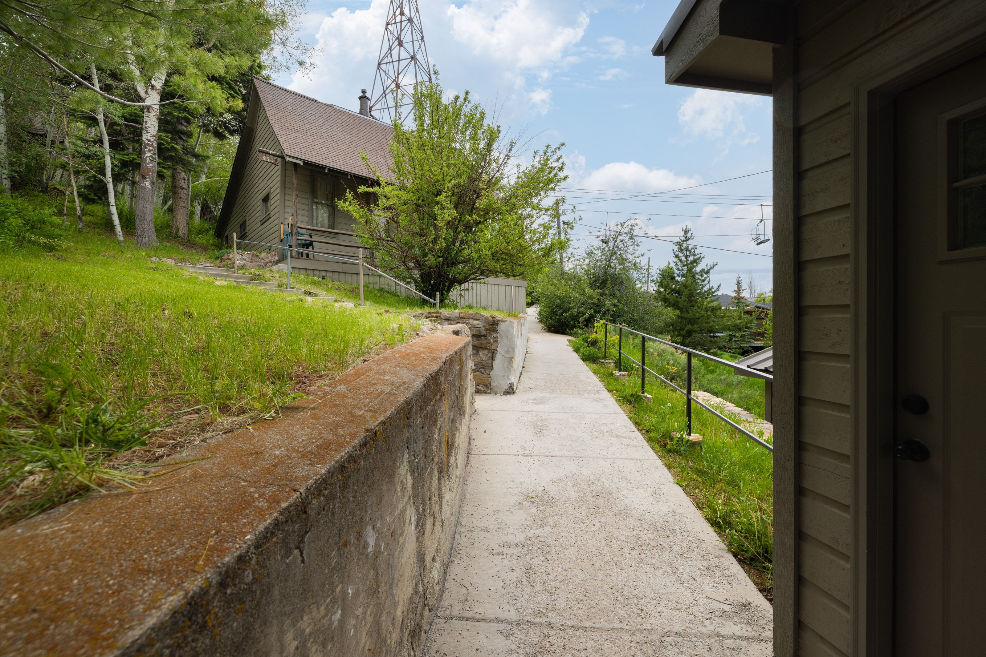 Concrete pathway with metal handrail alongside a retaining wall, leading toward a gabled dwelling with wood siding and a covered porch