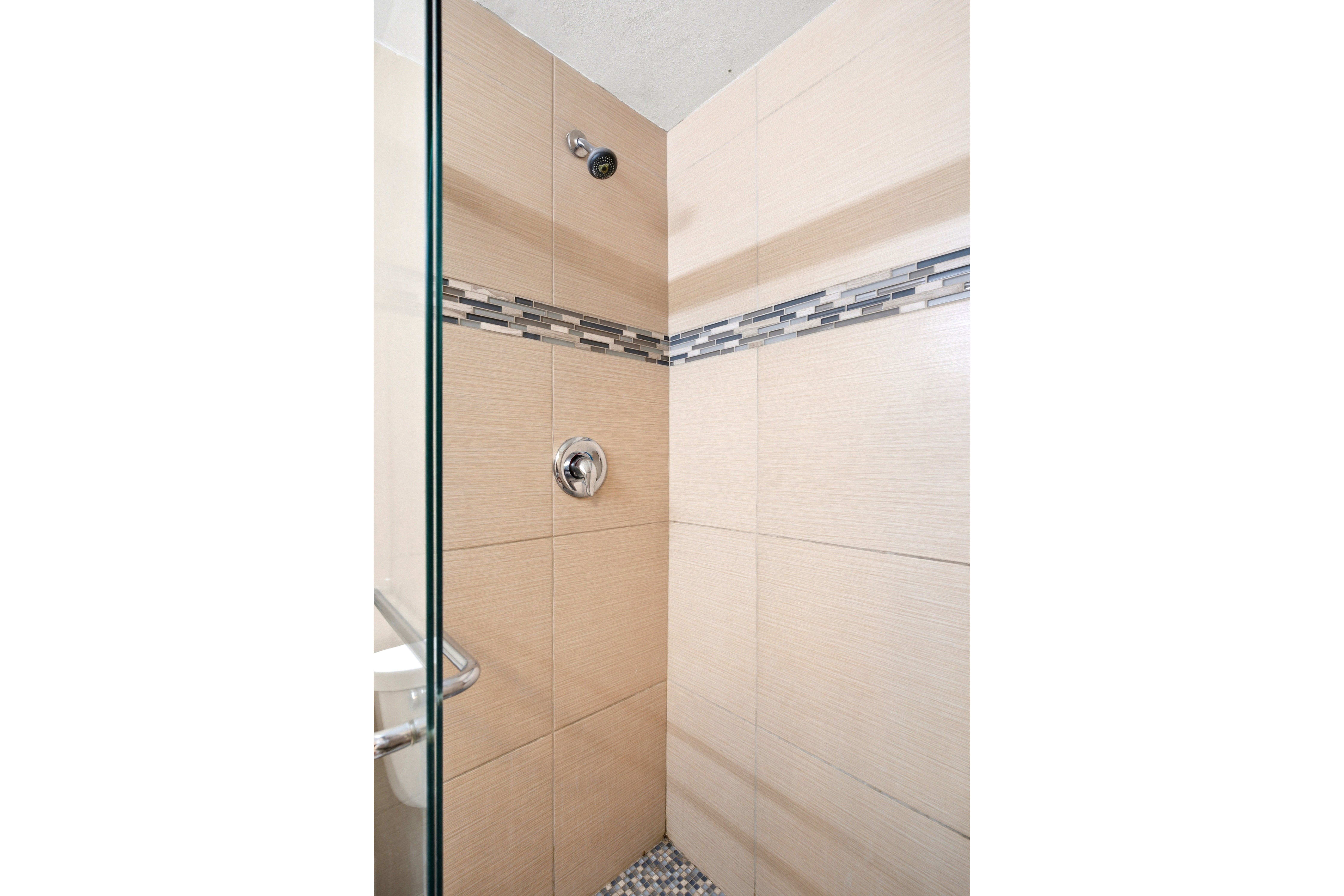 Shower enclosure featuring textured beige wall tiles, a mosaic accent band, pebble flooring, a glass panel, and polished chrome fixtures