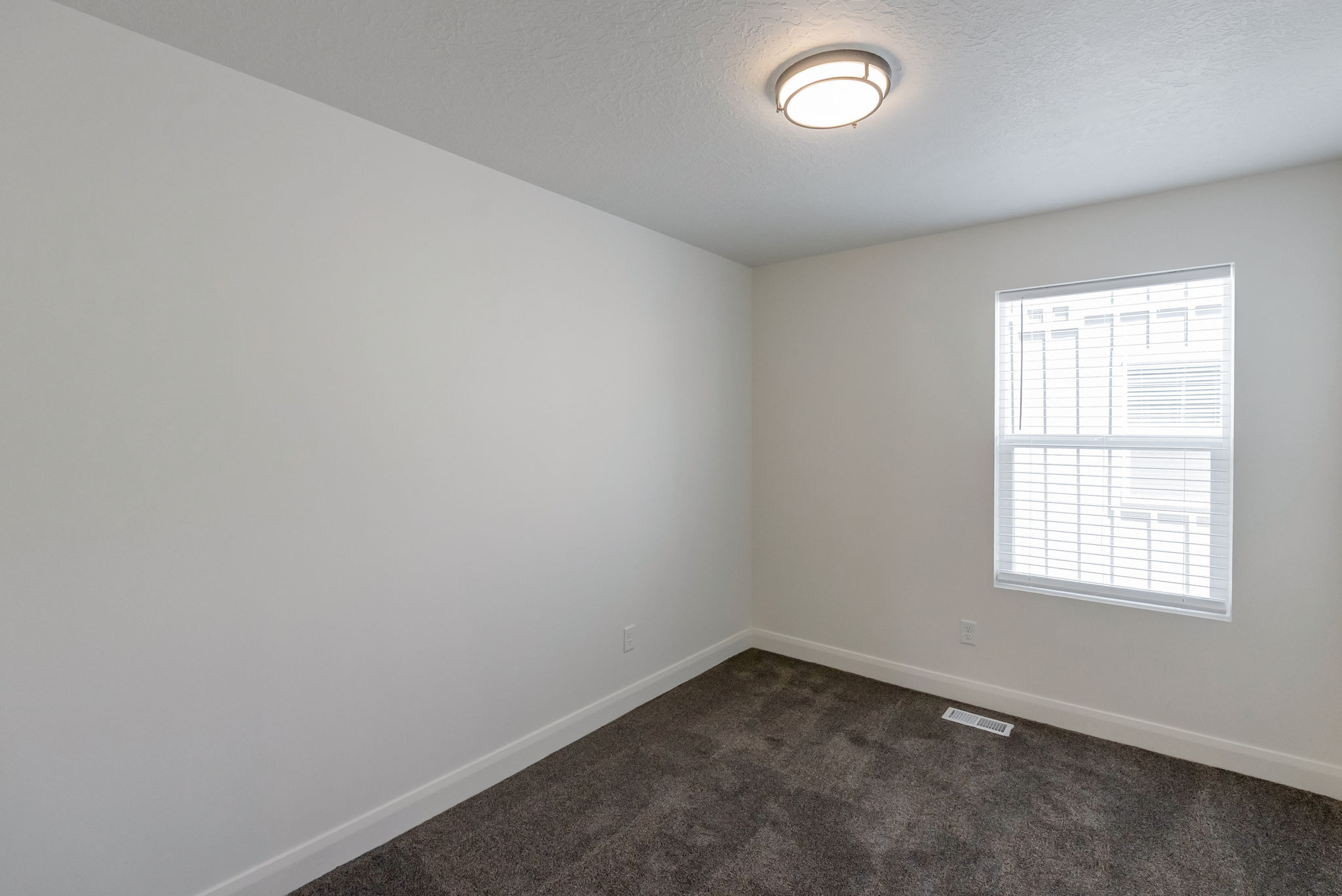 Neutral-toned room featuring a single window with blinds, a flush-mount ceiling light fixture, plush dark gray carpeting, and white baseboards