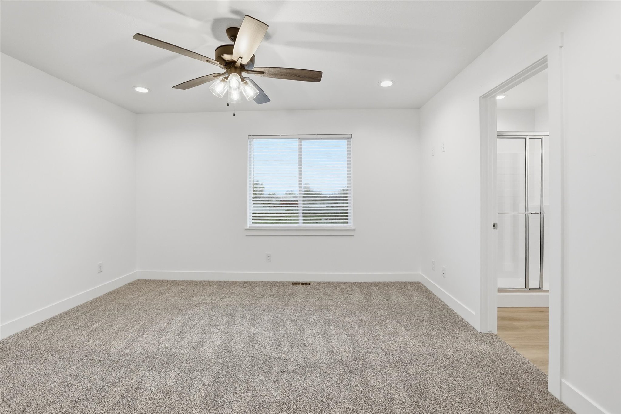 Carpeted interior space featuring a window with horizontal blinds, a ceiling fan with integrated lighting, recessed lighting, and a doorway opening to a bathroom with a glass-enclosed shower and wood-finish flooring
