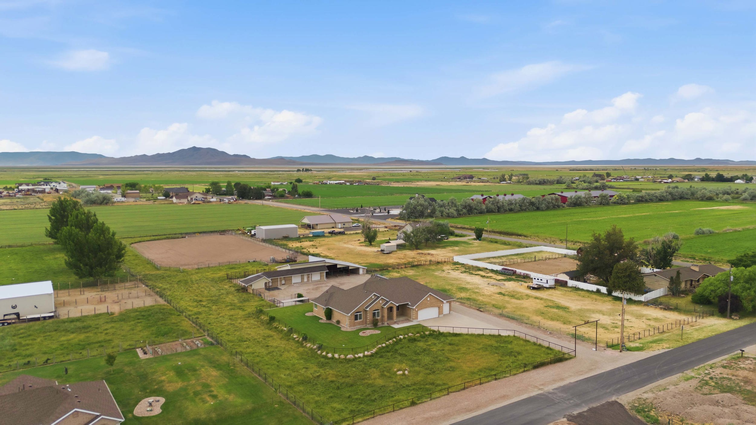 Expansive rural property featuring a tan stucco home with an attached garage, multiple outbuildings, fenced paddocks, and extensive green fields