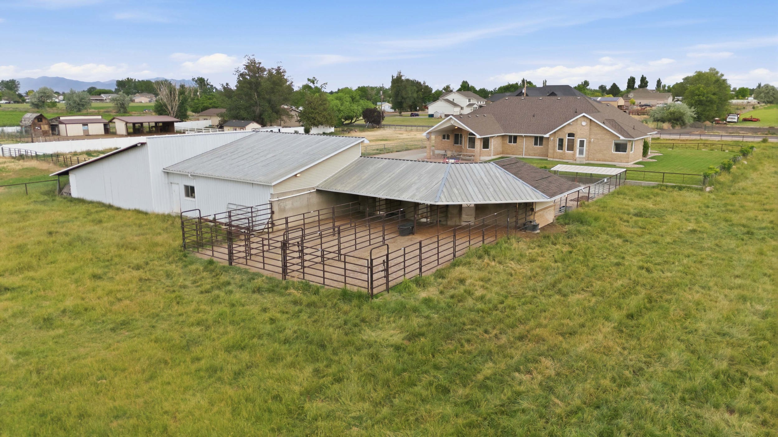 Expansive property featuring a brick exterior residence with a brown shingle roof, accompanied by a large white metal barn with a pitched roof, and a covered animal enclosure with metal fencing