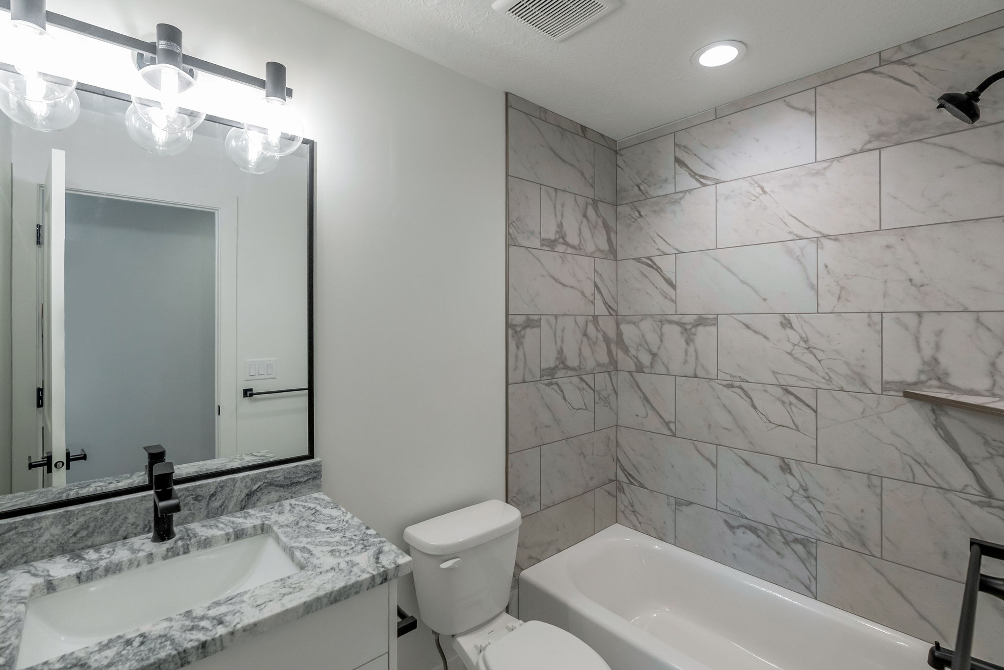 Bathroom featuring a white vanity with a patterned countertop, a matte black faucet, a large framed mirror, a bathtub with a tiled surround, and recessed lighting