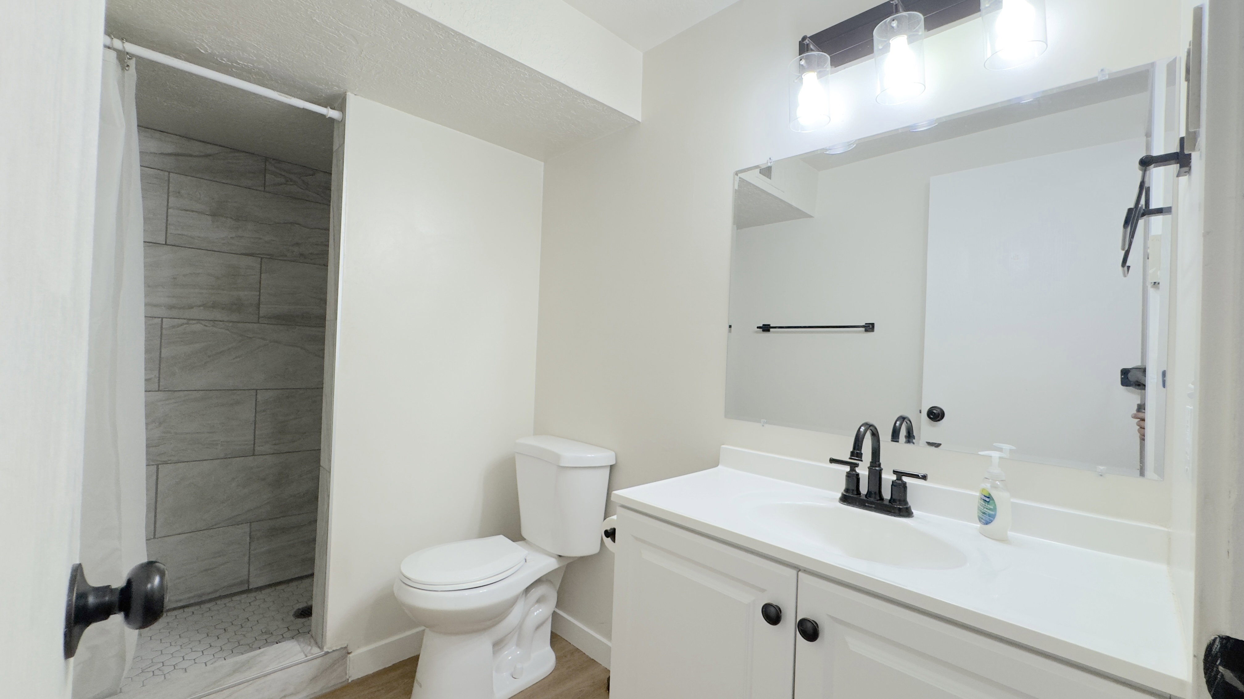 Hall way 3/4 bathroom featuring a white vanity with a single basin, a large wall-mounted mirror, and a three-light fixture with exposed bulbs.