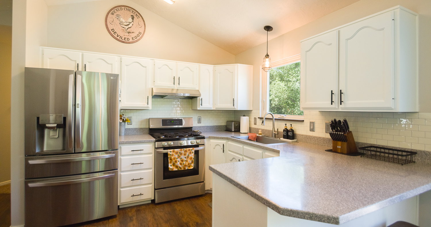 Kitchen featuring vaulted ceilings, white cabinetry with black hardware, stainless steel appliances, wood-finish flooring, and light-toned countertops