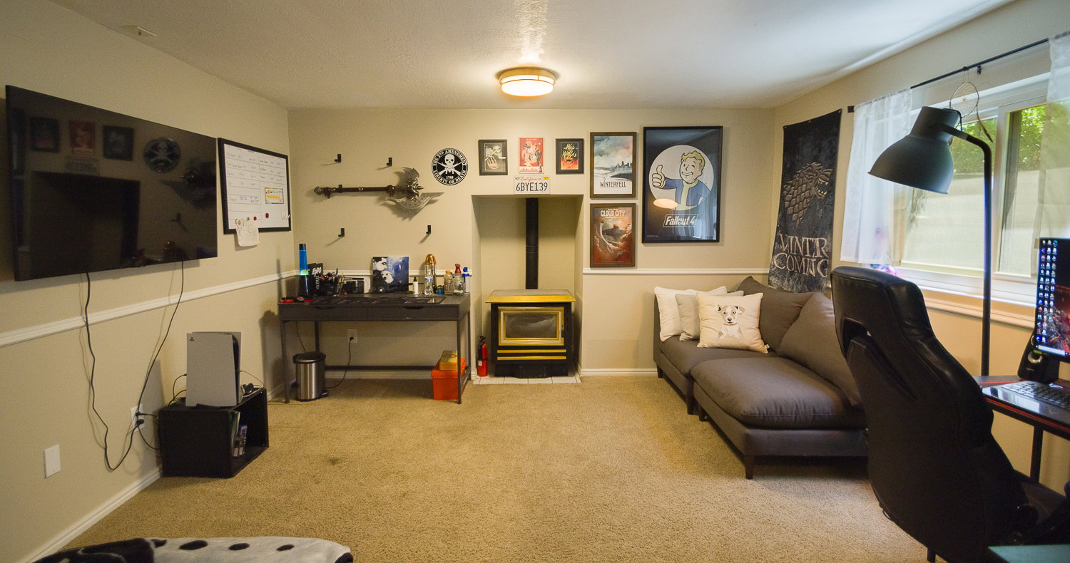 Carpeted recreation room featuring a built-in wood-burning stove with a stone hearth, wall-mounted television, and a window with white sheer curtains