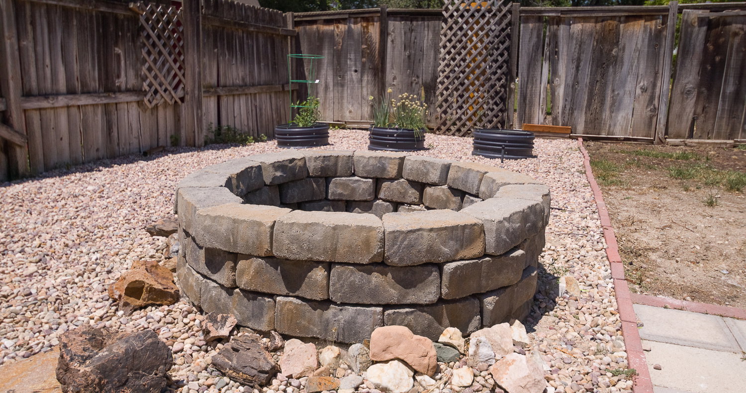 Backyard featuring a stone fire pit, gravel landscaping, a wooden privacy fence, and multiple planting areas