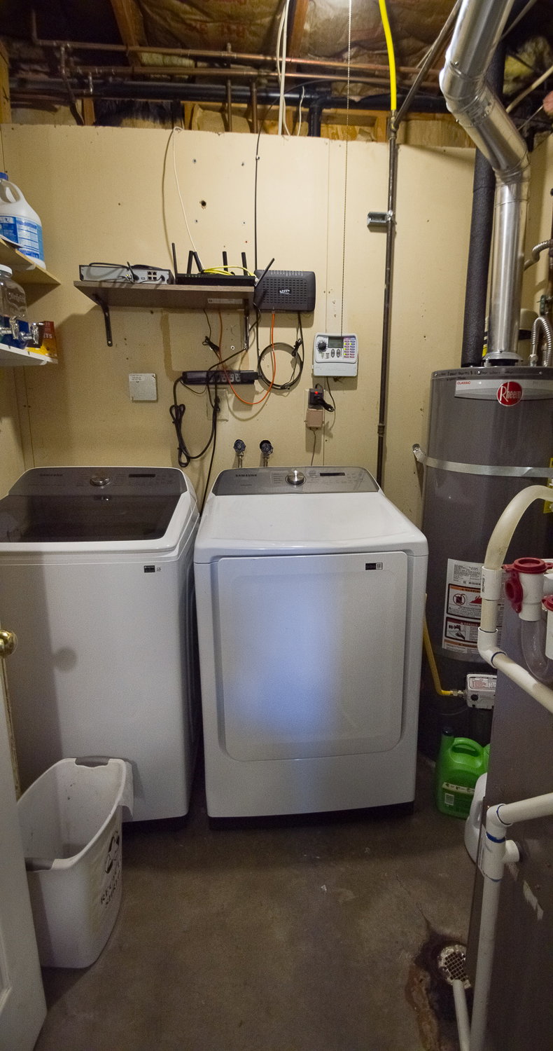 Utility area featuring a water heater, exposed overhead piping, concrete flooring, and a wall-mounted shelving unit