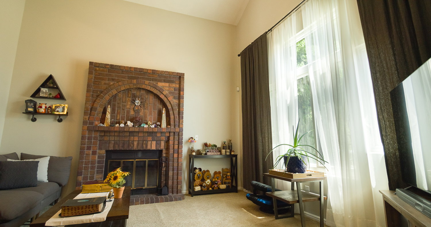 Living area featuring a brick fireplace with an arched design, light beige walls, vaulted ceilings, and a tall window with sheer white and dark brown drapery