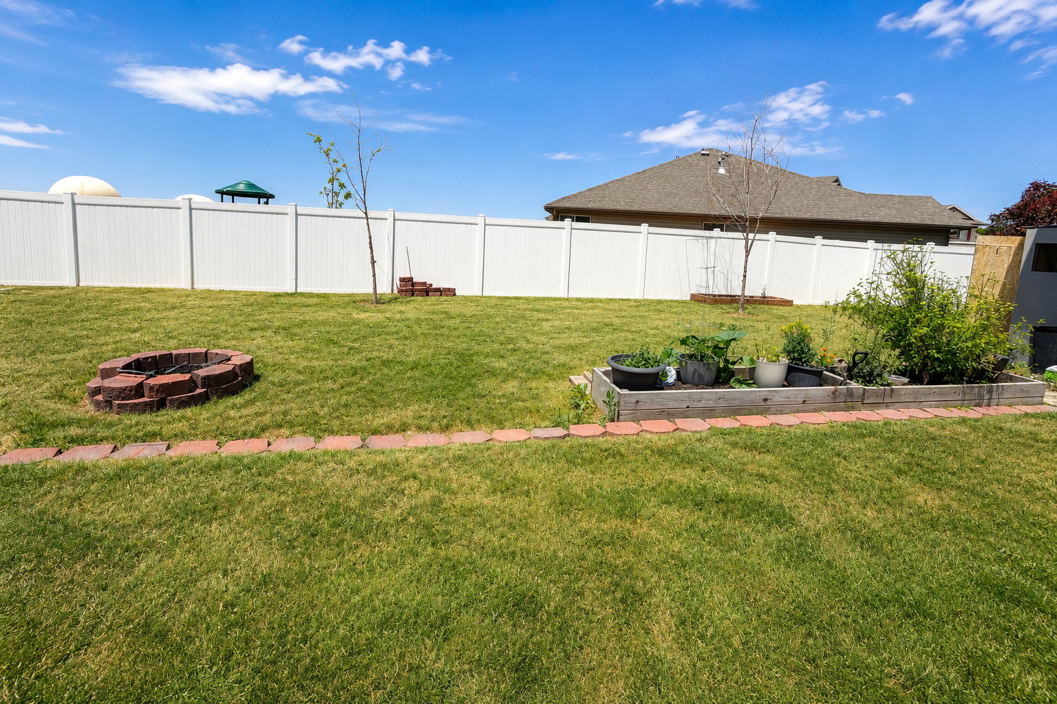 Expansive backyard featuring a lush lawn, white vinyl privacy fencing, and a brick paver fire pit