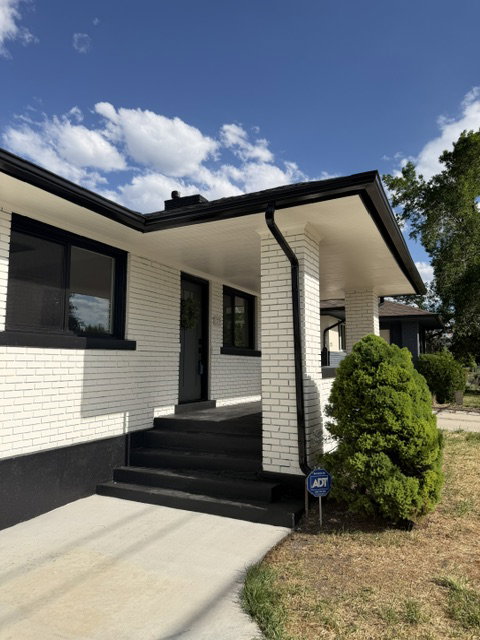 White brick exterior with black trim, a covered porch, black exterior steps, black gutters, and a compact evergreen shrub