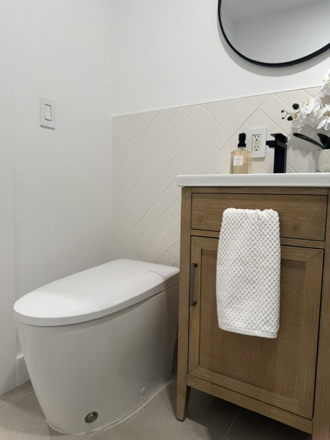 Modern bathroom featuring a contemporary toilet, wood-finish vanity with a woven panel door, matte black faucet, and white herringbone-patterned tile wainscoting