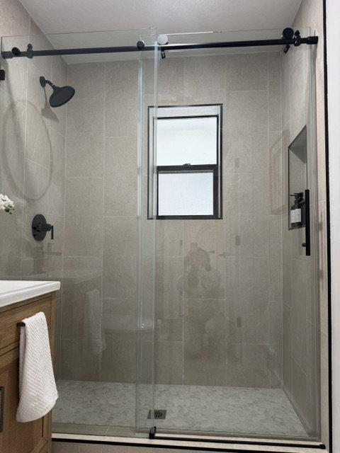 Enclosed shower featuring clear glass doors, matte black fixtures, light-toned wall tile, a window, and a pebble-finish shower floor