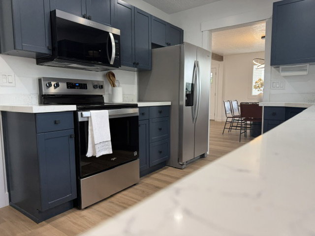 Modern kitchen featuring dark blue cabinetry, stainless steel appliances, white countertops, and wood-finish flooring