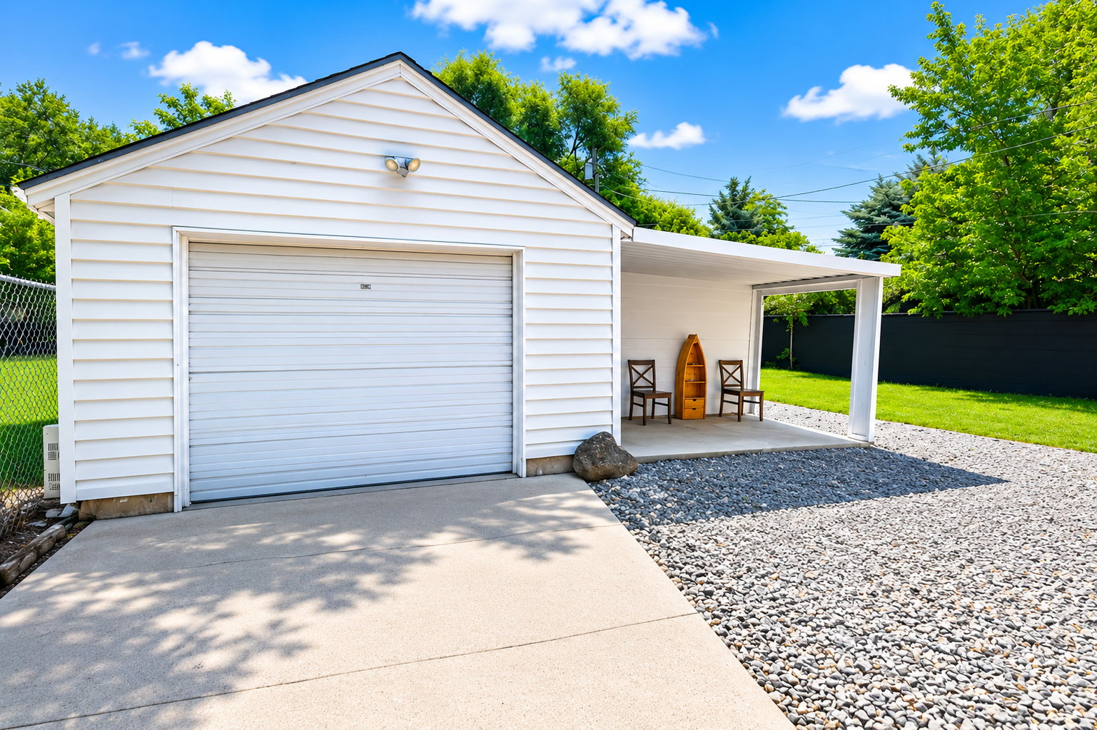 Detached single-car garage with white siding, concrete driveway, gravel parking area, and an attached covered carport