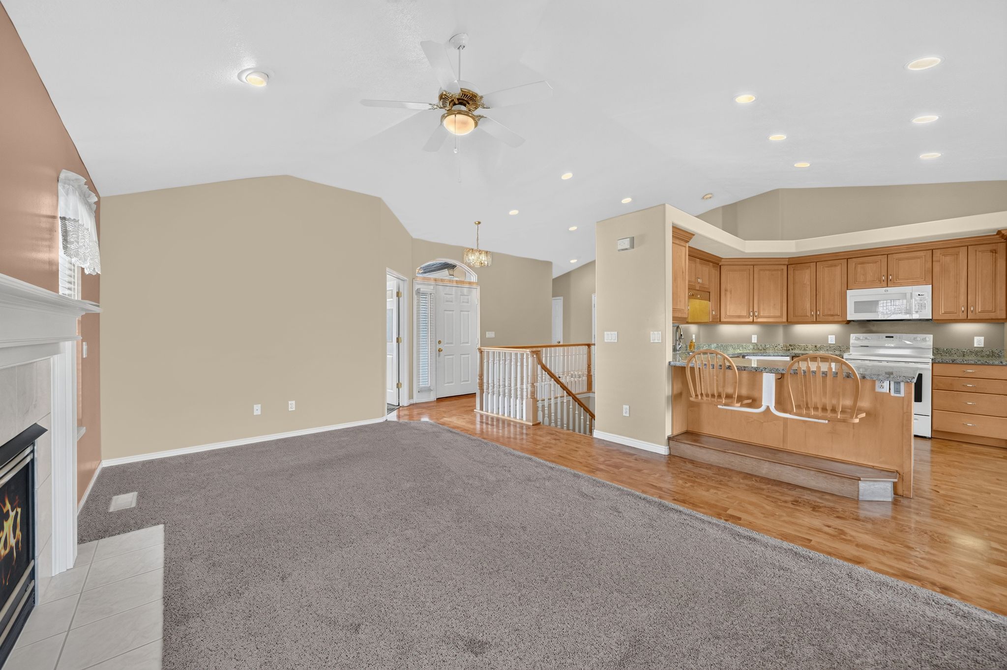 Open-concept living area featuring vaulted ceilings, a fireplace with a white mantle, wood-finish flooring, a kitchen with wood cabinetry and stainless steel appliances, and recessed lighting