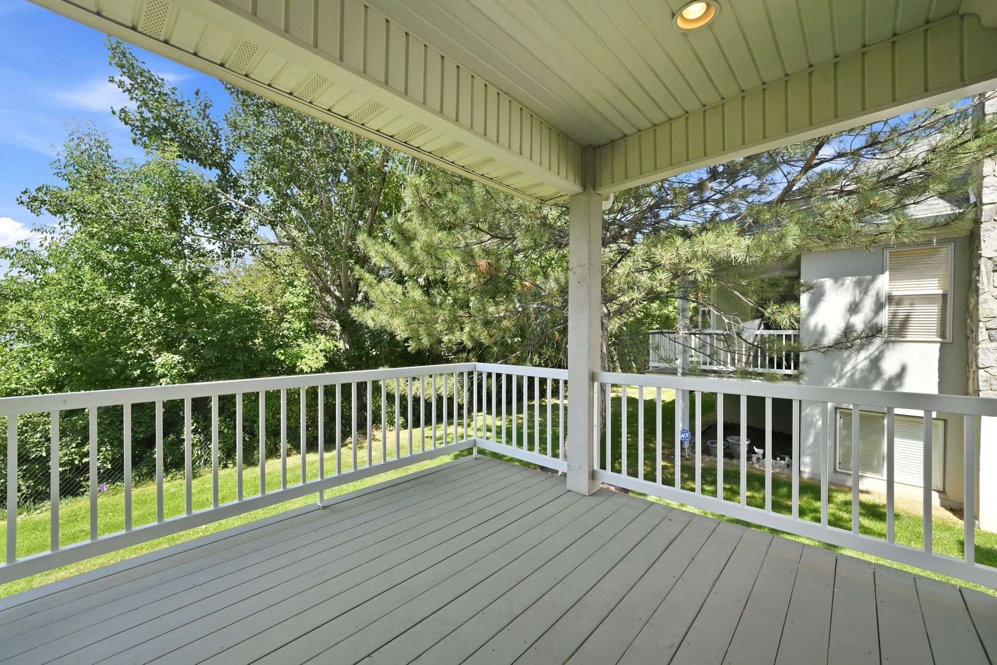 Covered outdoor deck with light gray wood-finish flooring, white railing, and a textured overhead ceiling featuring a recessed light fixture