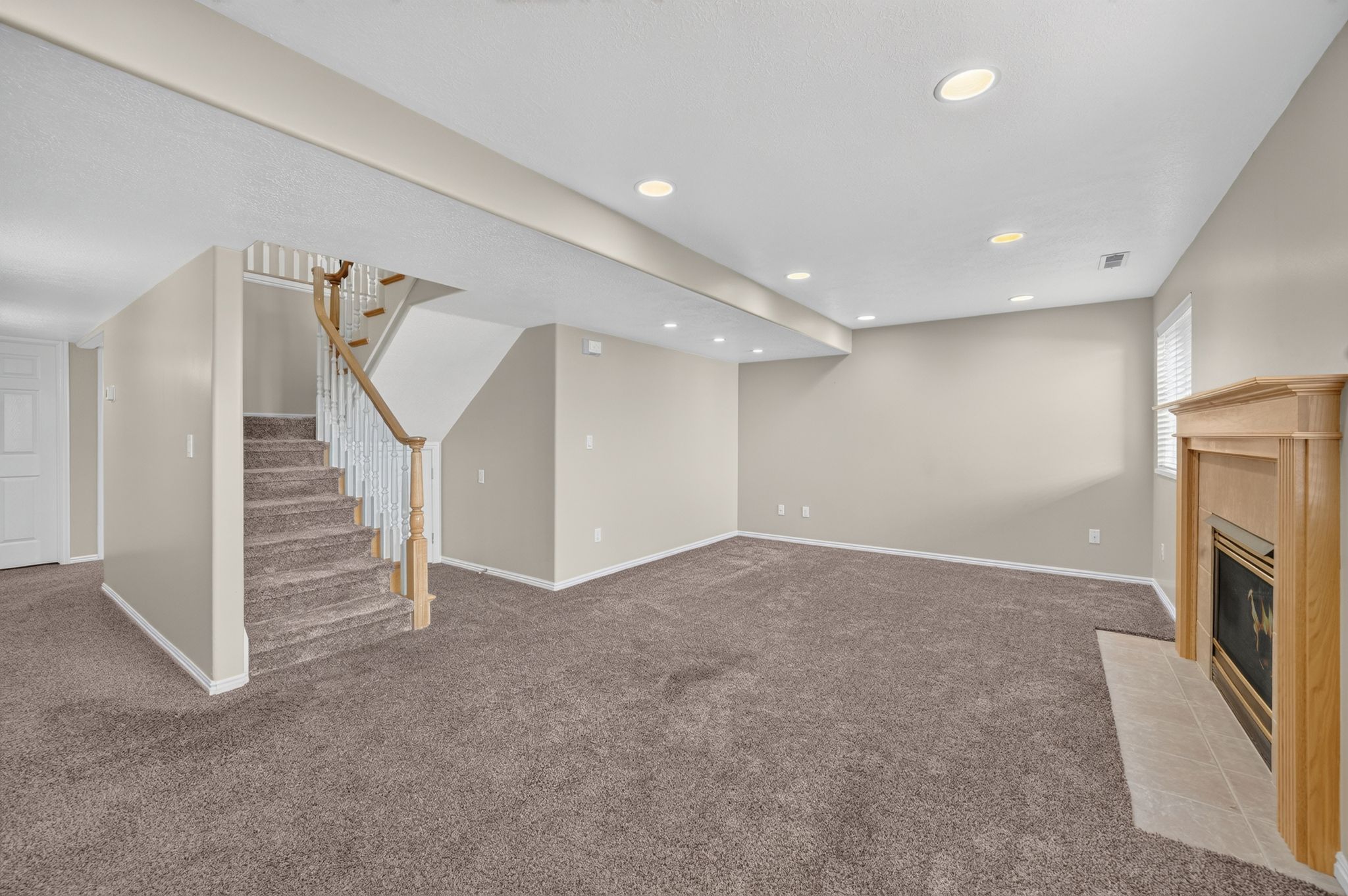 Spacious room featuring neutral carpeting, recessed lighting, a wood-surround fireplace with a tiled hearth, and a carpeted staircase with a wood handrail and white balusters